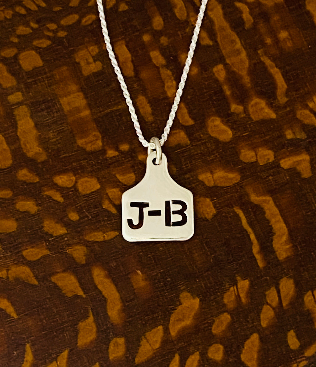 Mini sterling silver ear tag necklace with J-B initials on a ball chain, on cowhide