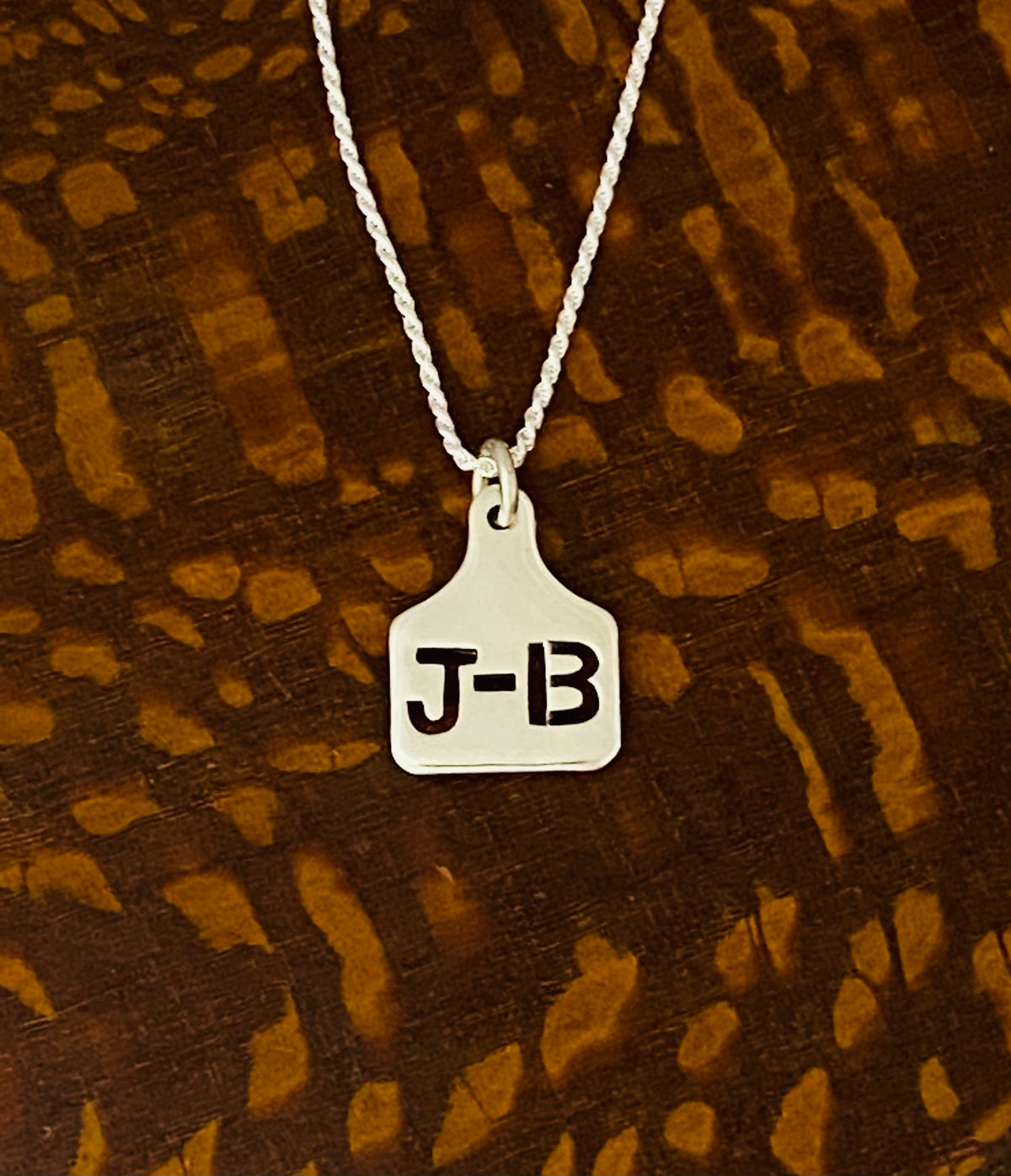 Mini sterling silver ear tag necklace with J-B initials on a ball chain, on cowhide